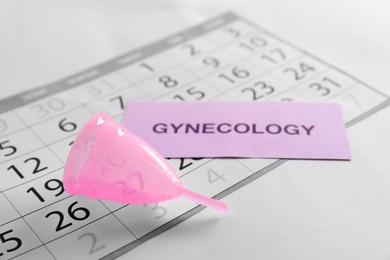 Menstrual cup, calendar and card with word GYNECOLOGY on light background