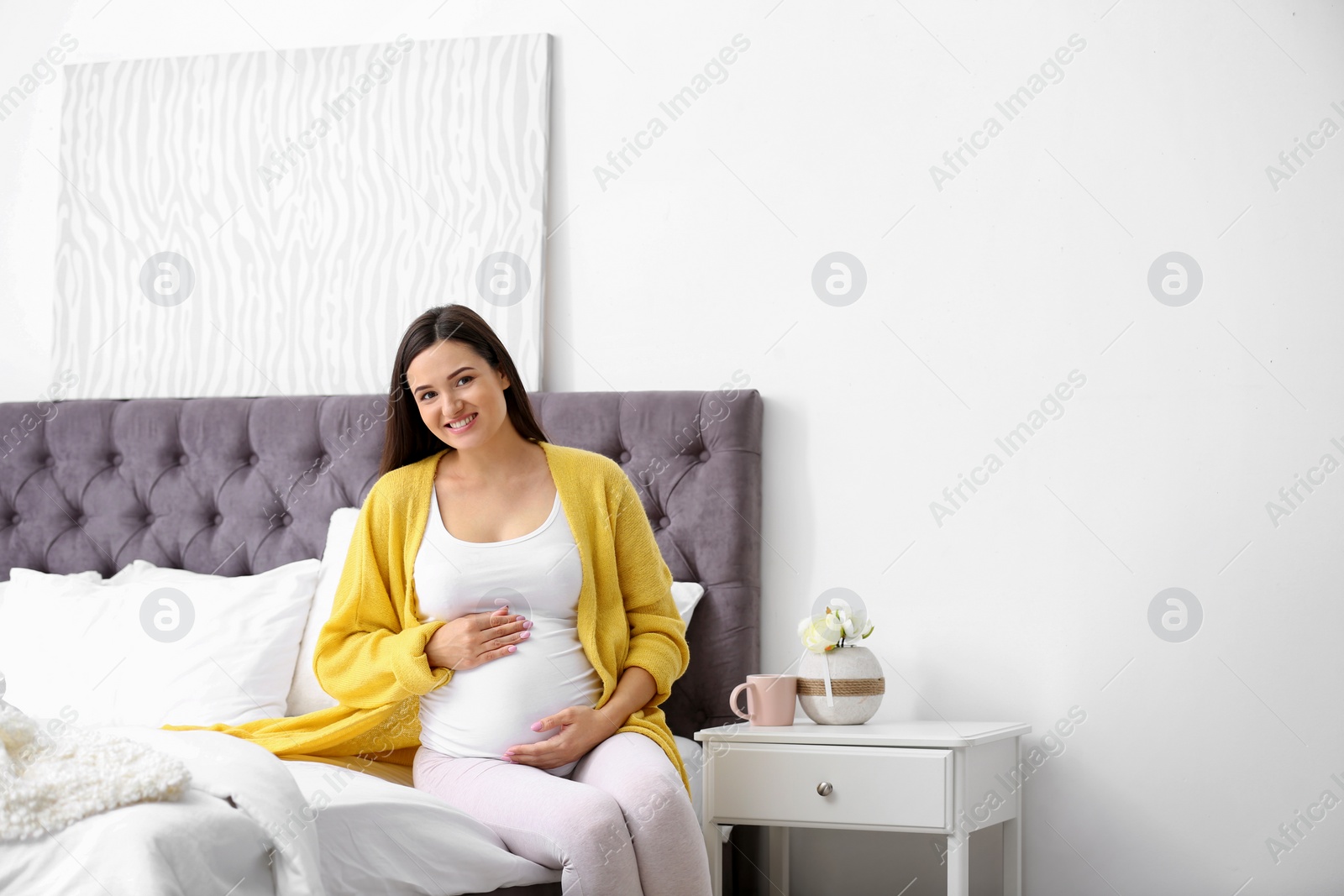 Photo of Young beautiful pregnant woman sitting on bed and touching her belly at home