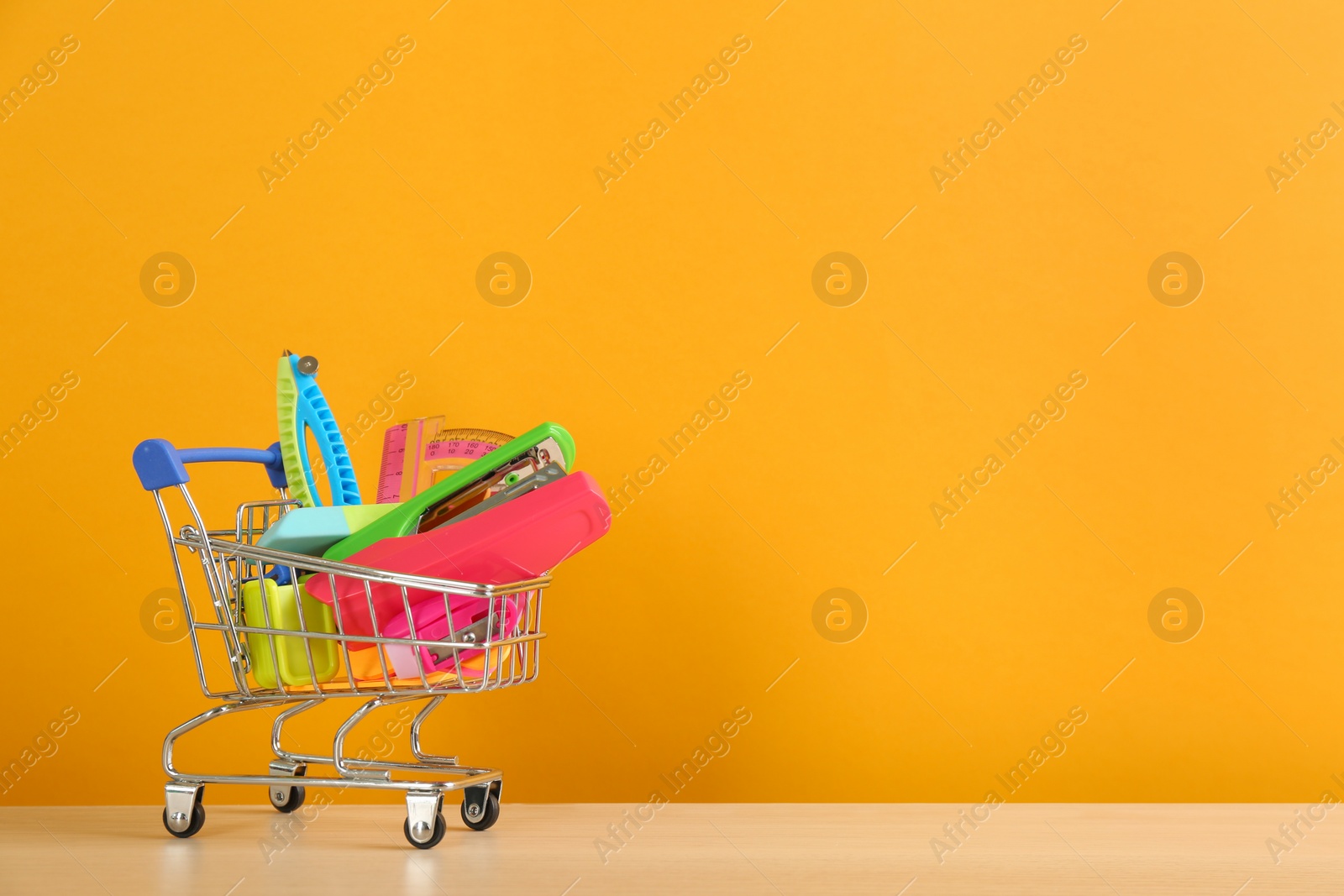 Photo of Different school stationery in miniature shopping cart on table against orange background, space for text. Back to school
