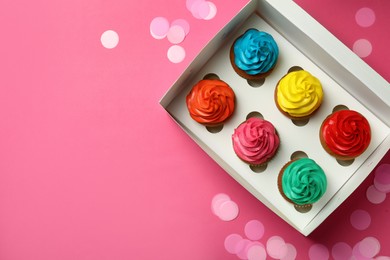 Box with different cupcakes and confetti on pink background, flat lay. Space for text