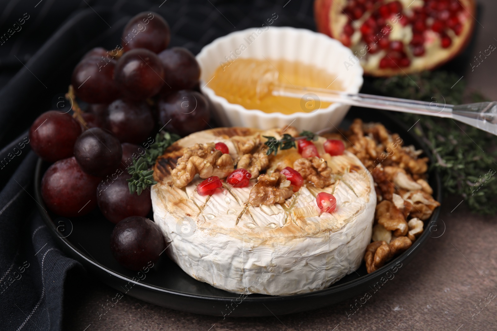 Photo of Plate with tasty baked camembert, honey, grapes, walnuts and pomegranate seeds on brown table, closeup