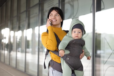 Photo of Mother holding her child in sling (baby carrier) while talking on smartphone near building outdoors