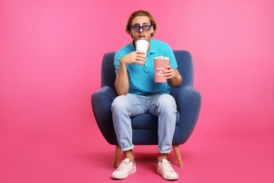 Emotional man with 3D glasses, popcorn and beverage sitting in armchair during cinema show on color background