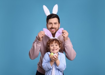 Photo of Happy father and son in cute bunny ears having fun on light blue background. Easter celebration