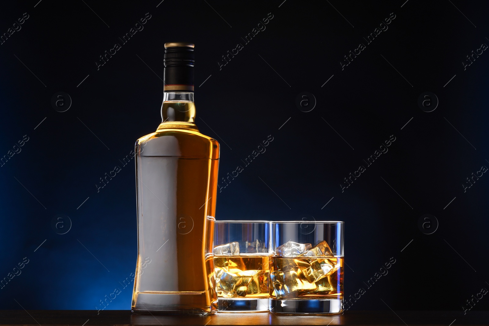 Photo of Whiskey with ice cubes in glasses and bottle on table against dark background