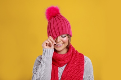 Photo of Funny young woman in warm sweater, scarf and hat on yellow background. Winter season