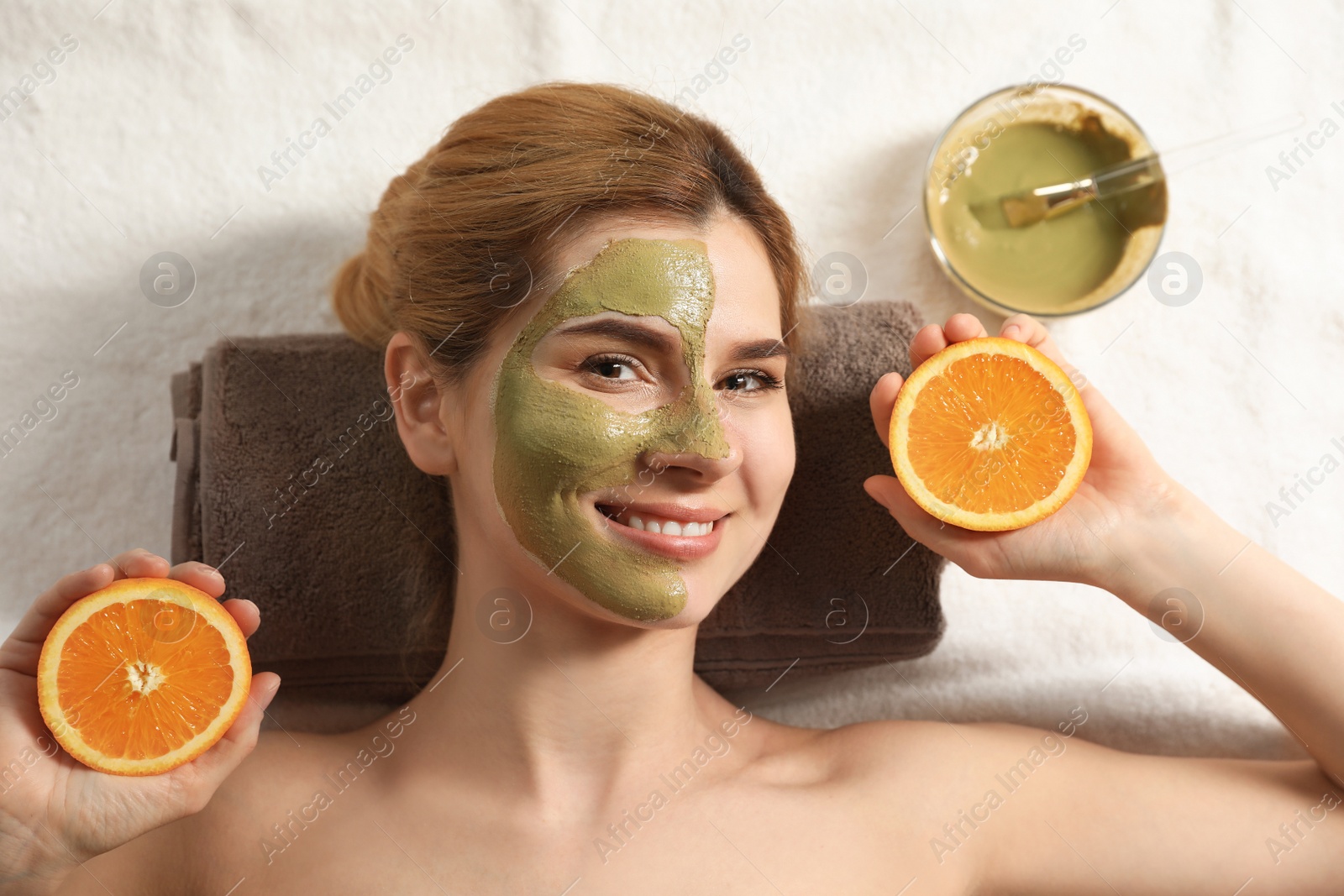 Photo of Beautiful woman with mask on face and cut orange relaxing in spa salon, top view