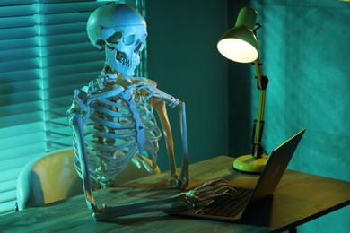 Photo of Waiting concept. Human skeleton sitting at wooden table with laptop indoors