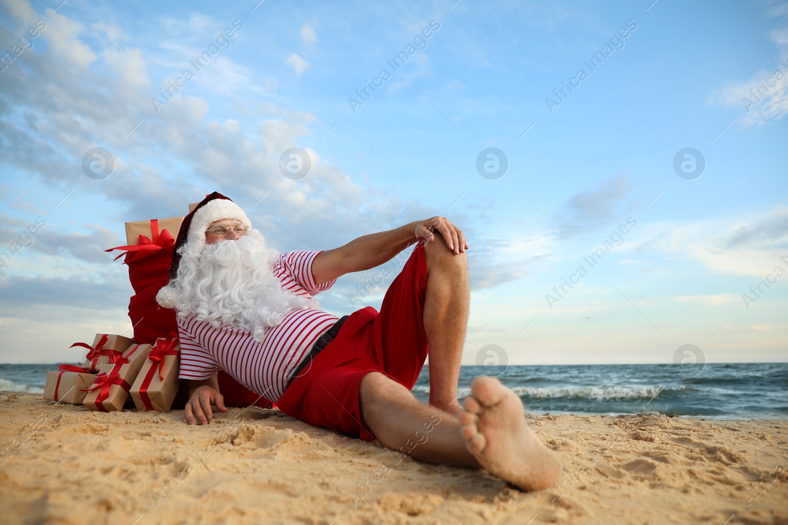 Photo of Santa Claus with bag of presents relaxing on beach, space for text. Christmas vacation