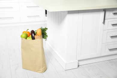 Photo of Paper shopping bag full of vegetables and baguette on floor in kitchen. Space for text