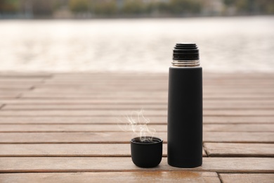 Photo of Black thermos and cap on wooden pier near river. Space for text