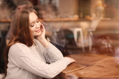 Photo of Beautiful young woman sitting at table in cafe, view from outdoors through window
