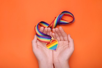 Photo of Woman holding rainbow ribbon with heart shaped pendant on orange background, top view. LGBT pride