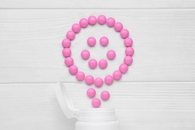 Photo of Happy emoticon made of pink antidepressants and medical jar on white wooden table, flat lay