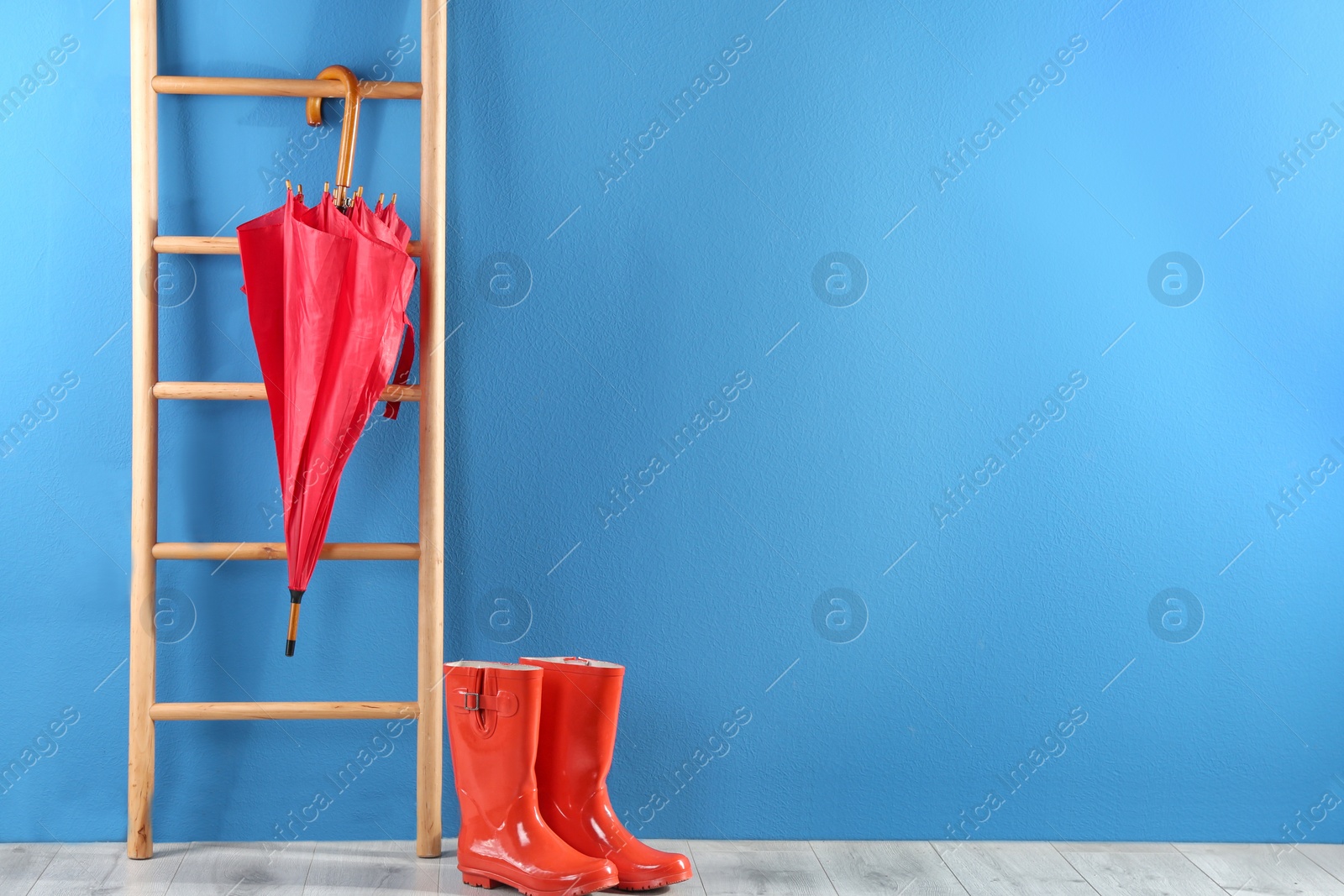 Photo of Beautiful red umbrella on ladder and rubber boots near blue wall. Space for text