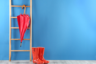 Photo of Beautiful red umbrella on ladder and rubber boots near blue wall. Space for text