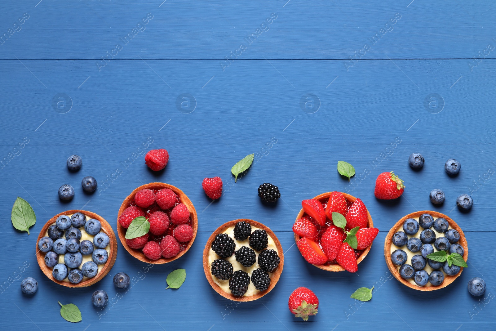 Photo of Tartlets with different fresh berries on blue wooden table, flat lay and space for text. Delicious dessert