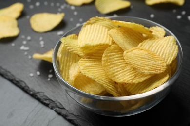 Photo of Slate board with bowl of potato chips on grey table, closeup. Space for text