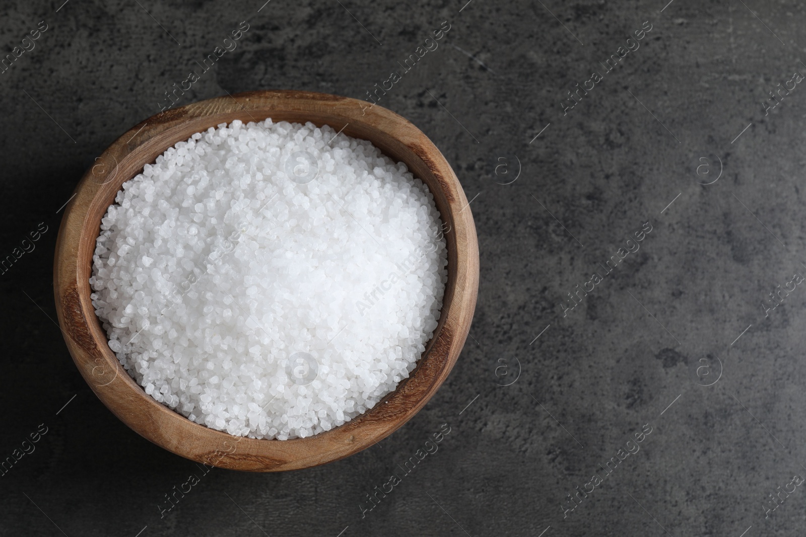 Photo of Natural salt in wooden bowl on dark grey table, top view. Space for text