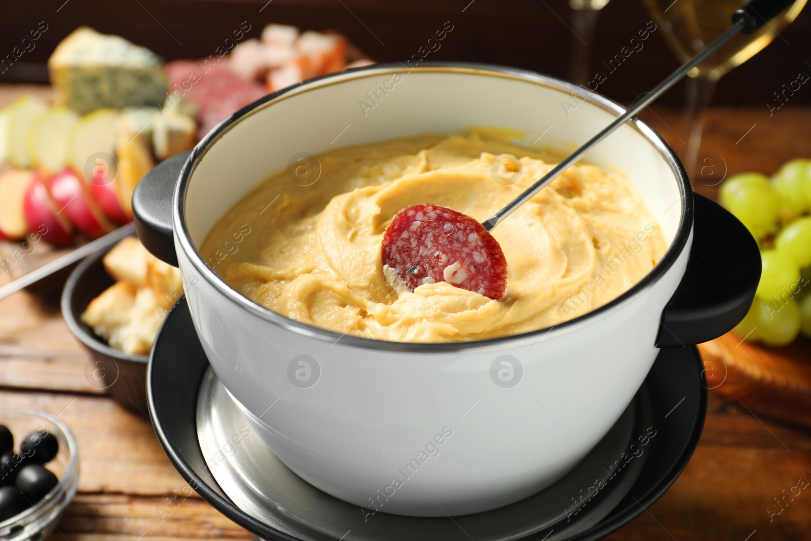 Photo of Fork with piece of sausage and melted cheese in fondue pot on wooden table, closeup