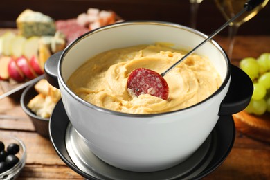 Photo of Fork with piece of sausage and melted cheese in fondue pot on wooden table, closeup