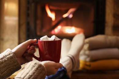 Woman with cup of hot drink resting near fireplace indoors, closeup. Space for text