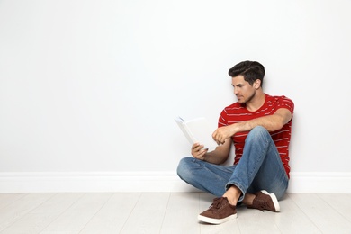 Handsome man reading book on floor near white wall, space for text