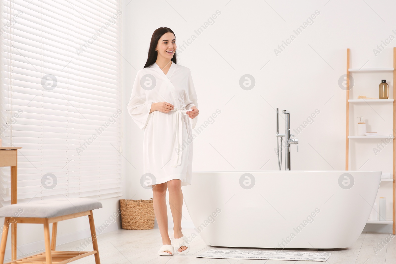 Photo of Beautiful happy woman wearing stylish bathrobe near tub in bathroom