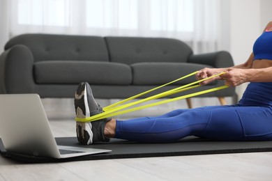 Photo of Woman doing exercise with fitness elastic band near laptop on mat at home, closeup