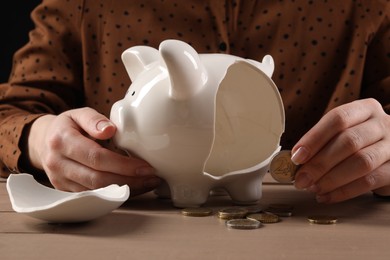 Poverty. Woman holding coin near broken piggy bank at wooden table, closeup
