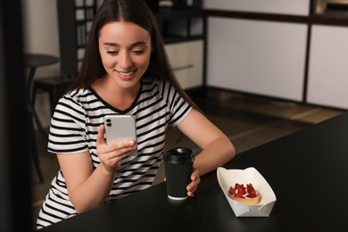 Photo of Happy young woman with paper cup of coffee and smartphone at table in hostel dining room