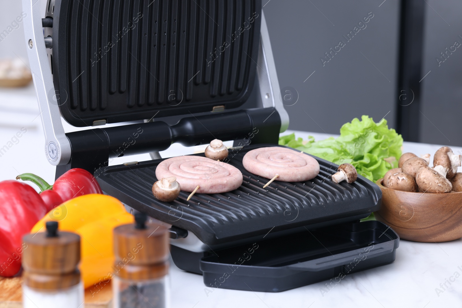 Photo of Electric grill with homemade sausages, mushrooms, and vegetables on white table indoors