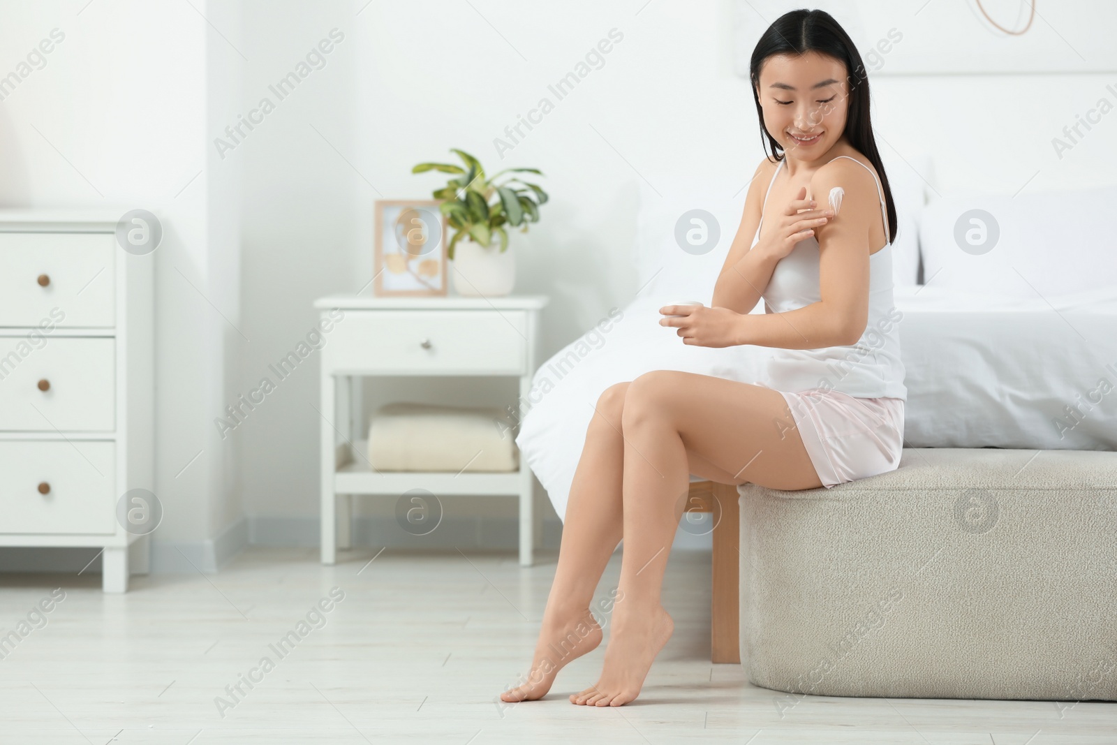 Photo of Beautiful young Asian woman applying body cream on shoulder in bedroom, space for text
