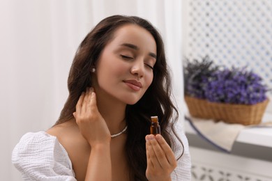 Beautiful young woman applying essential oil onto neck indoors