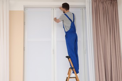 Worker in uniform installing roller window blind on stepladder indoors