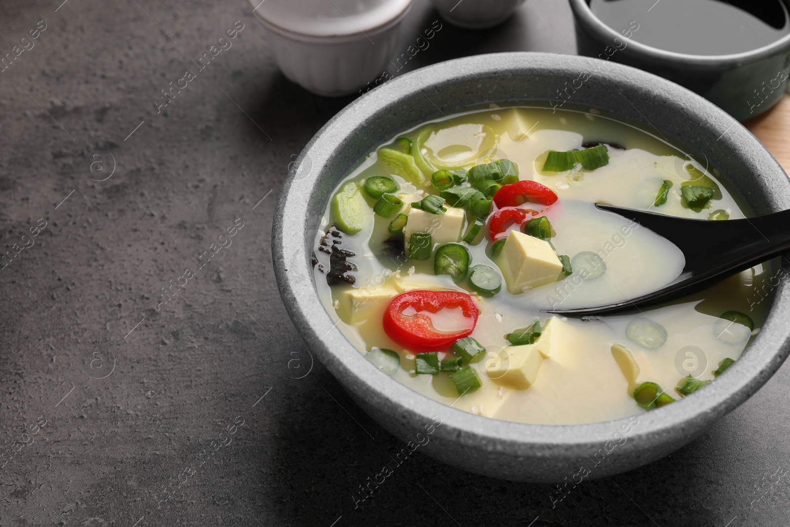 Photo of Bowl of delicious miso soup with tofu and spoon served on grey table. Space for text