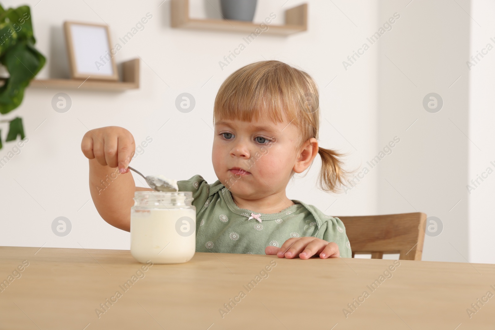 Photo of Cute little child eating tasty yogurt with spoon from jar at wooden table