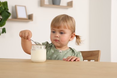 Cute little child eating tasty yogurt with spoon from jar at wooden table