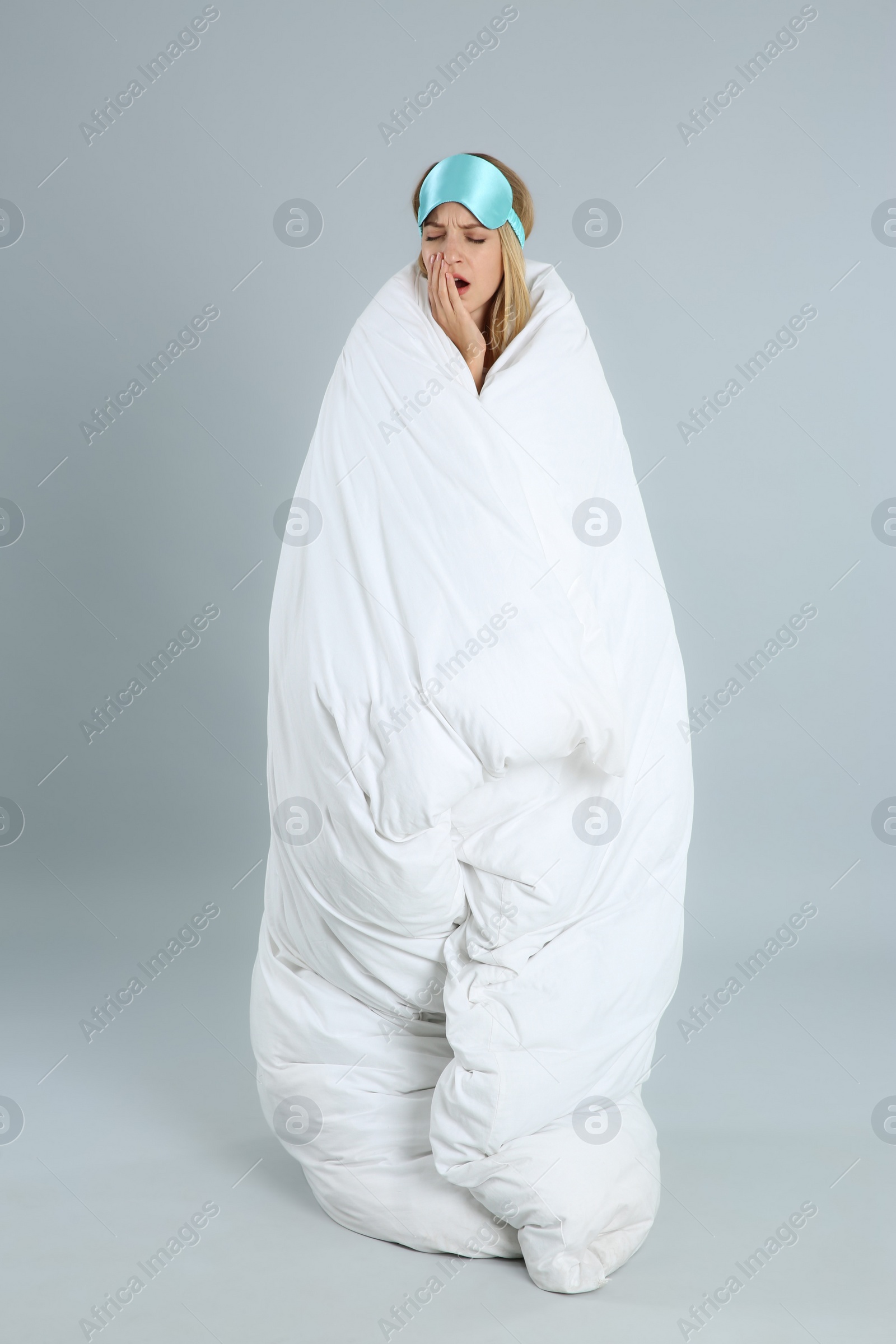 Photo of Young woman in sleeping mask wrapped with soft blanket yawning on grey background