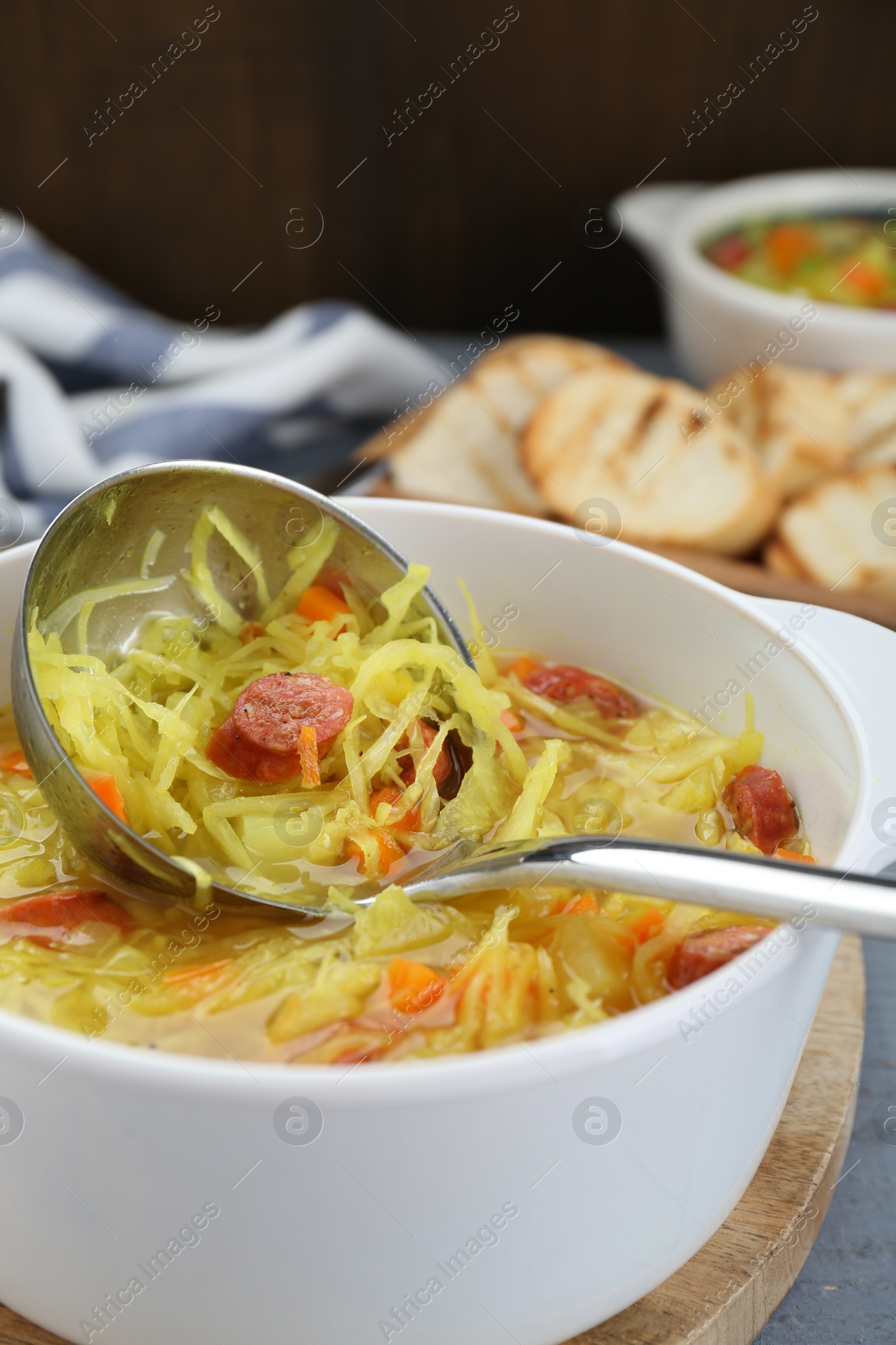 Photo of Bowl of delicious sauerkraut soup with smoked sausages and ladle on board, closeup