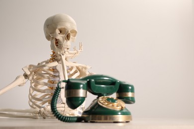 Waiting concept. Human skeleton at table with corded telephone against light grey background, space for text