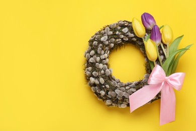 Wreath made of beautiful willow, colorful tulip flowers and pink bow on yellow background, top view. Space for text