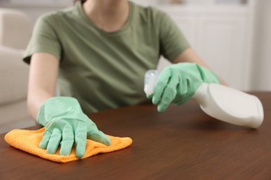 Photo of Woman with spray bottle and microfiber cloth cleaning wooden table in room, closeup