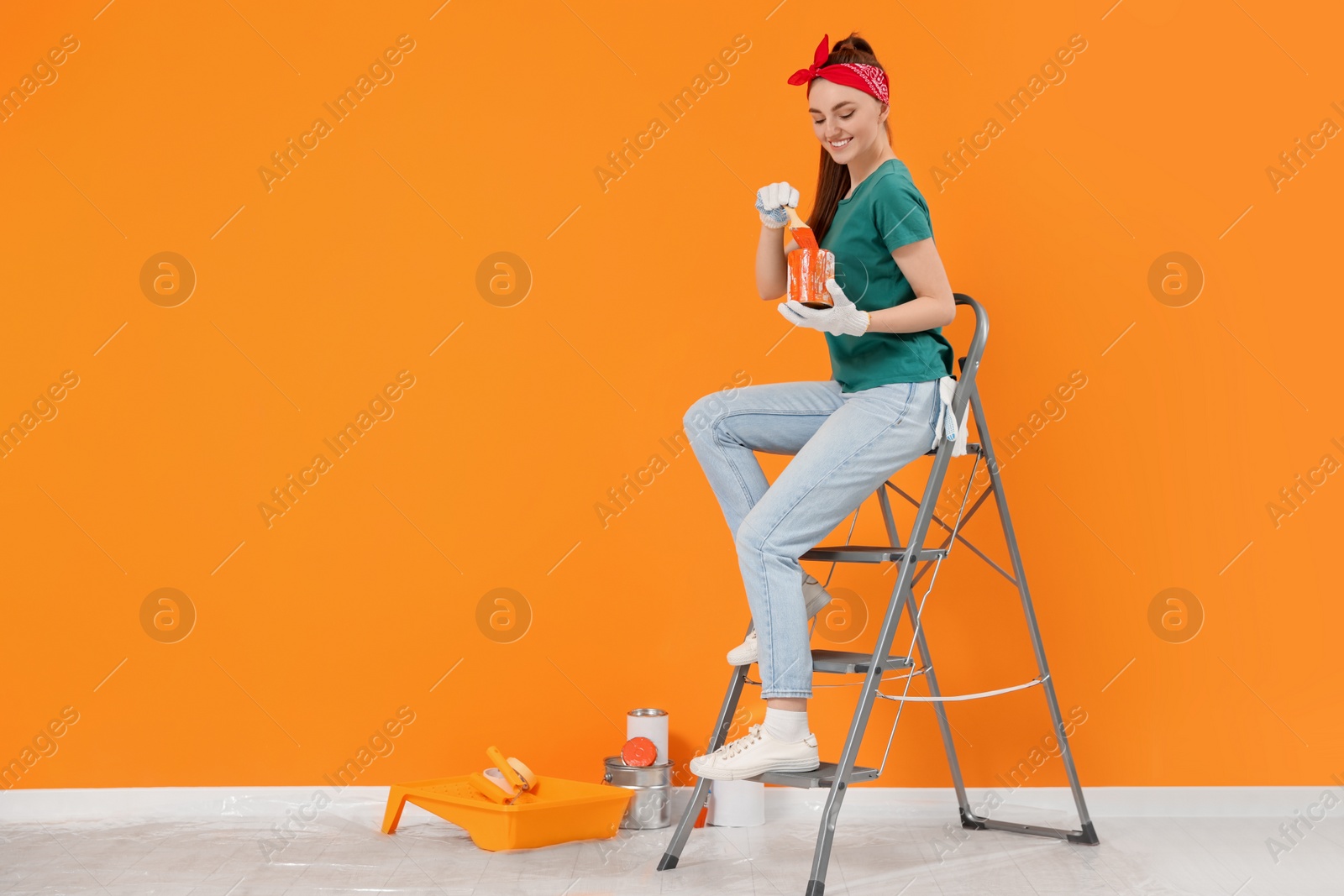 Photo of Happy designer with painting equipment sitting on folding ladder near freshly painted orange wall indoors, space for text