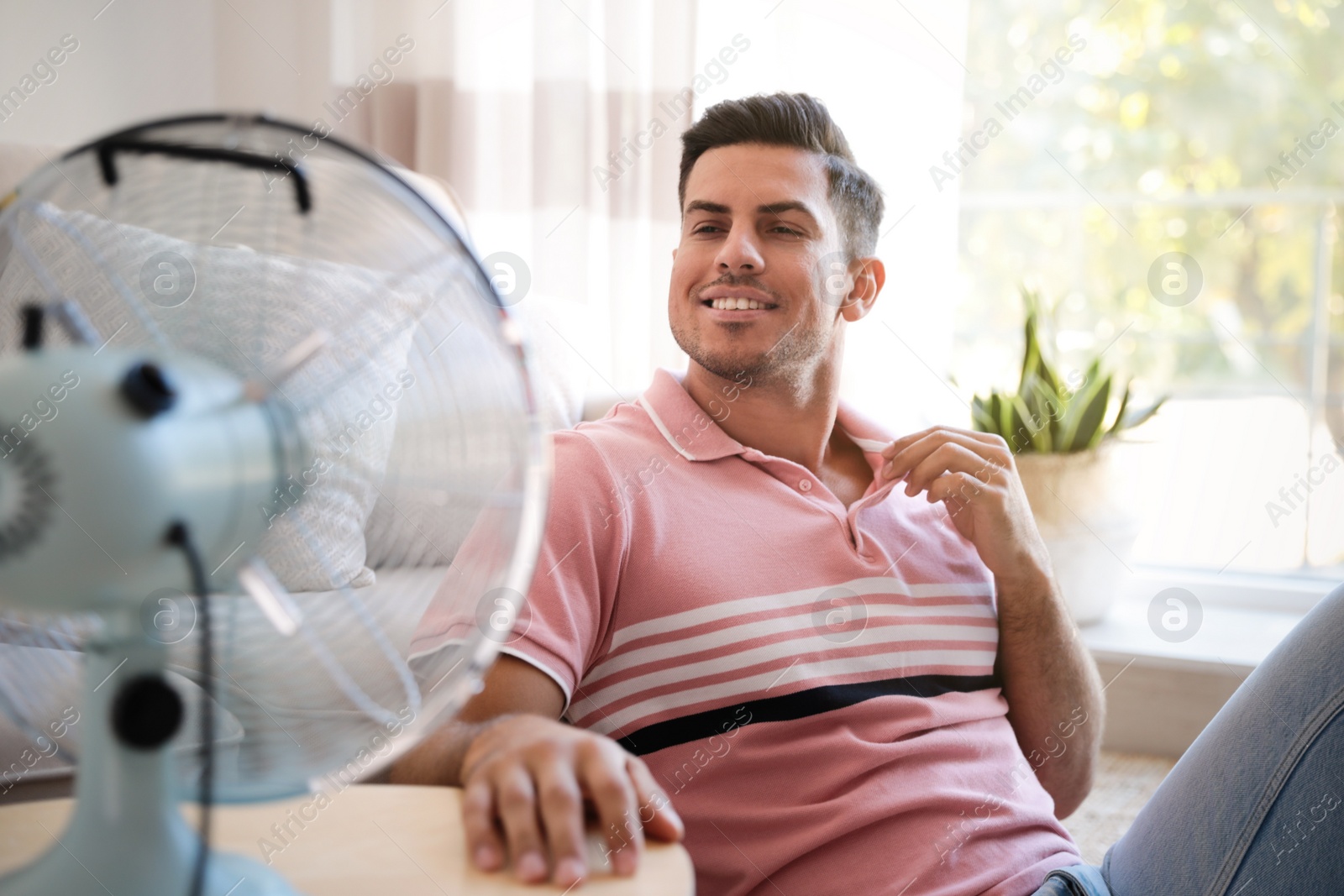 Photo of Man enjoying air flow from fan in living room. Summer heat