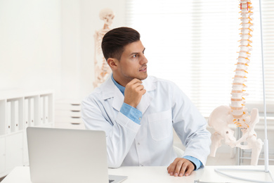 Photo of Male orthopedist with laptop near human spine model in office