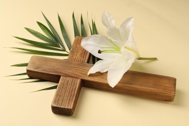 Wooden cross, lily flower and palm leaf on pale yellow background. Easter attributes