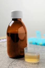 Photo of Bottle of syrup and measuring cup on light grey table. Cold medicine