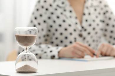 Photo of Hourglass with flowing sand on desk. Woman taking notes indoors, selective focus