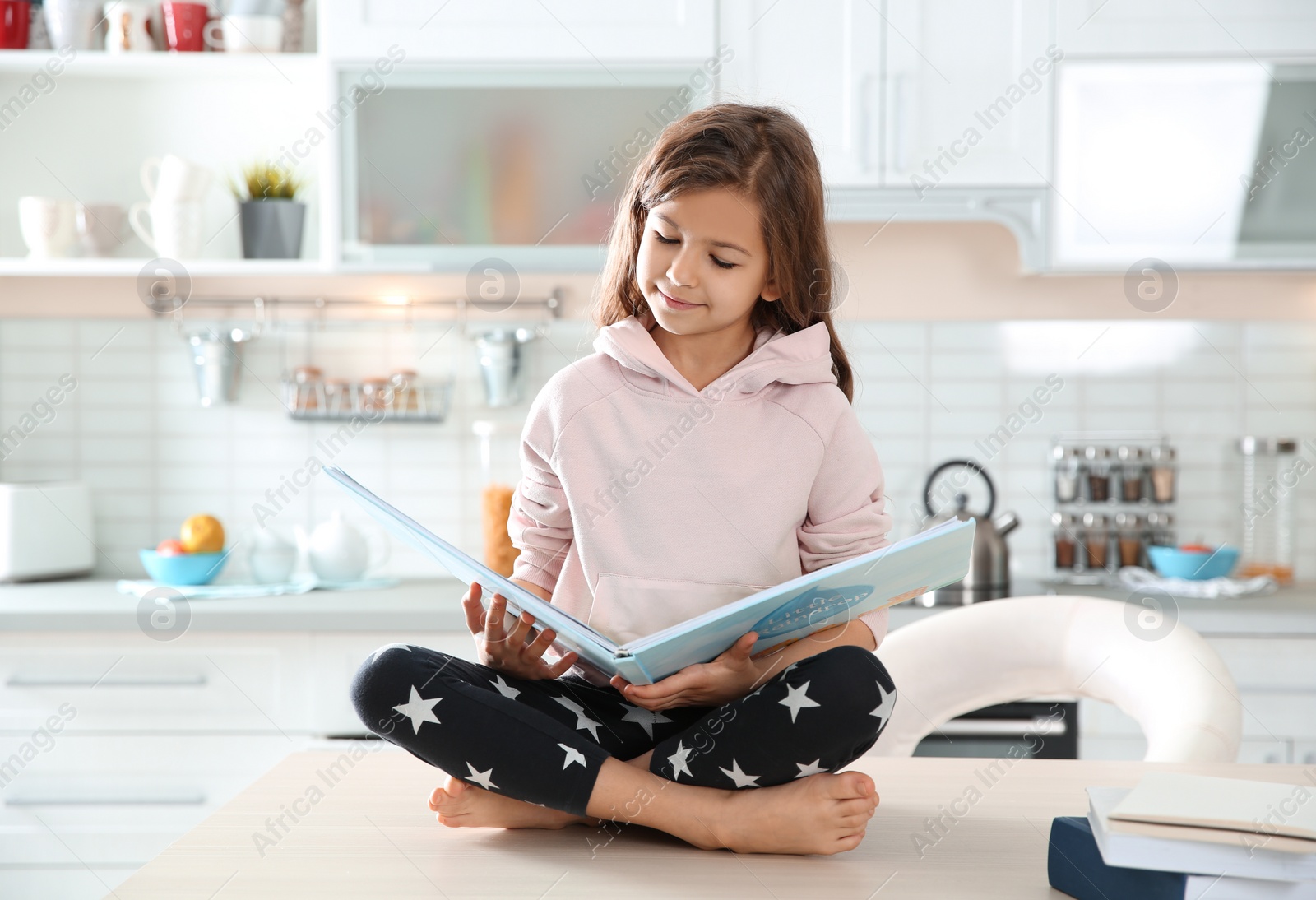 Photo of Cute little girl reading book in kitchen at home